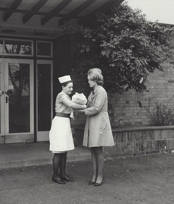 A black and white portrait photograph of a smiling nurse handing a newborn baby to its mother. They are stood outside the doorway to a building and are both dressed in 1960s-era clothing.