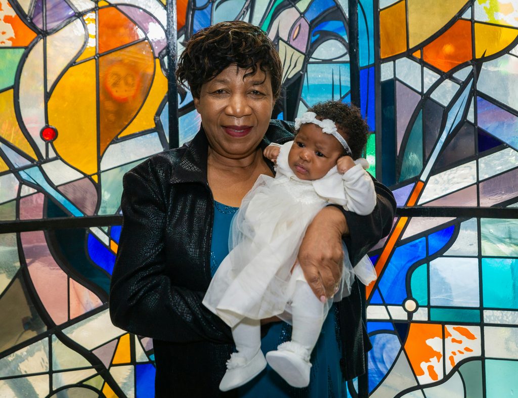 A 21st century photograph of an older Black woman holding a Black baby dressed in a white christening gown. They are stood in front of a vibrant, multi-coloured stained glass window.
