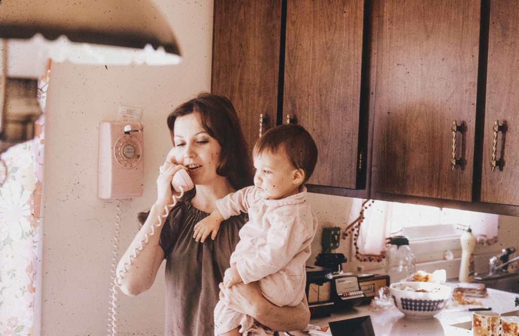 A 1970s photograph of a woman stood in her kitchen, holding her baby in one arm whilst making a phone call. The scene appears cosy and domestic.