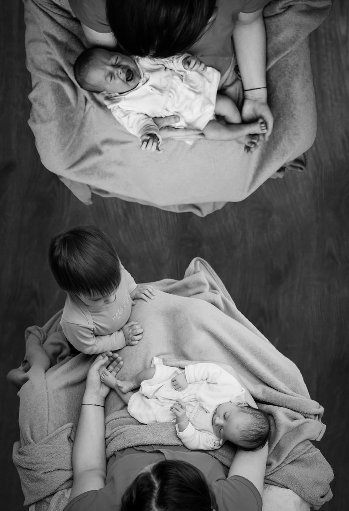 A black and white photograph of two women each holding a newborn baby in their laps. Next to one of the women a young toddler is stood gazing at the baby. 
